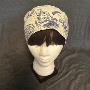 May include: A cream-colored surgical scrub cap with a blue toile pattern featuring a car, trees, and a bird. The cap is displayed on a mannequin head with brown hair.