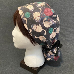 May include: A black scrub hat with a tie, featuring a repeating pattern of white cats wearing astronaut helmets and floating in space. The cats are holding yarn balls and surrounded by stars.