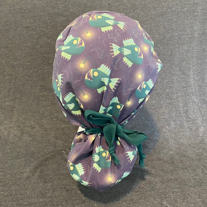 May include: A purple scrub hat with a green tie. The hat has a repeating pattern of anglerfish with glowing lures.