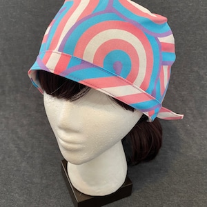May include: A colorful head covering with a swirling pattern in shades of pink, blue, white, and purple. The fabric is wrapped around a mannequin head, showcasing the design and fit. The head covering has a band around the edge.