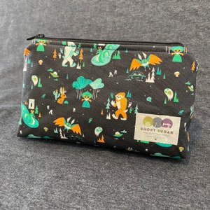 May include: Black zippered pouch with a whimsical forest pattern featuring Bigfoot, aliens, and other fantasy creatures. The pouch has a rectangular shape and a small label that reads "SHORT SUGAR FINE HANDMADE GOODS PROVIDENCE, RI."