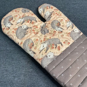 May include: A brown and beige oven mitt with a quilted pattern featuring a repeated design of opossums. The mitt has a brown and white checkered trim.