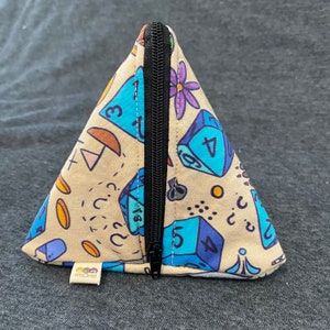 May include: A beige triangular zippered pouch with a black zipper. The pouch is decorated with a pattern of blue and white dice, flowers, and other geometric shapes.