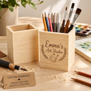 Puede incluir: Dos portalápices cuadrados de madera clara. Uno está en blanco, mientras que el otro presenta un grabado que dice Emma's Art Studio con un icono de paleta de pintura y pincel. Una pequeña etiqueta rectangular dice CUSTOM ENGRAVED.