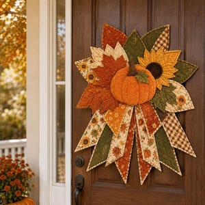 Puede incluir: Un tapiz de tela con una calabaza acolchada naranja central rodeada de hojas y tiras de tela superpuestas en tonos naranja, amarillo, marrón y verde. El diseño incluye patrones de girasoles, cuadros y texturas acolchadas.