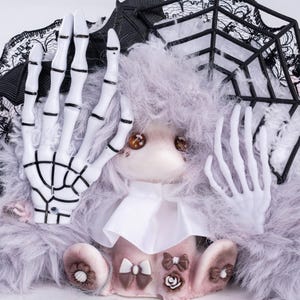 Könnte beinhalten: Eine Plüschpuppe mit langem, zotteligem hellgrauem Fell und zwei weißen Kunststoffskeletthänden an den Seiten des Gesichts. Die Puppe hat braune Knopfaugen, einen weißen Stoffkragen sowie kleine braune Schleifen und Rosenverzierungen an den Füßen.