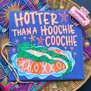 Könnte beinhalten: Blaues T-Shirt mit dem Text "Hotter Than a Hoochie Coochie" und einer Grafik eines grünen Alligators, der auf einem Tie-Dye-Schwimmring schwimmt. Das Design umfasst Palmen und Sterne. Eine rosa-weiß karierte Haarspange und eine Perlenkette sind ebenfalls sichtbar.
