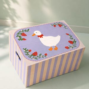 Puede incluir: Caja de almacenamiento de madera con exterior a rayas morado claro y beige. La tapa presenta un ganso blanco con un lazo rojo, rodeado de fresas y hojas verdes sobre un fondo morado claro. La caja tiene un asa lateral.