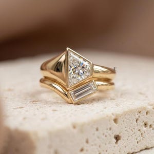 May include: A gold ring set consisting of two bands. The top band features a large triangular-cut diamond, while the bottom band is curved and set with a rectangular baguette-cut diamond. Both bands are crafted from polished yellow gold metal.