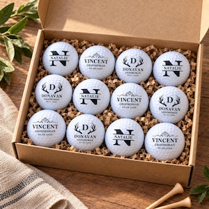 May include: A set of twelve white golf balls arranged in a cardboard box filled with brown paper filler. Each ball features black printed text and graphics, including the names Natalie, Vincent, or Donavan, along with dates and antler motifs.