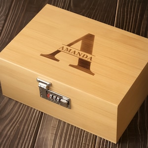 Könnte beinhalten: Eine rechteckige, helle Holzbox mit einem silberfarbenen Zahlenschloss an der Vorderseite. Die Oberseite ist mit einem großen Buchstaben A graviert, in dessen Mitte der Name AMANDA in dunkelbrauner Farbe horizontal eingearbeitet ist.