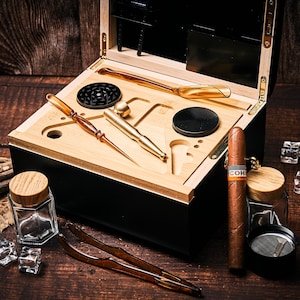 May include: A black wooden box with a light wood interior containing a gold-coloured metal pipe, a black grinder, a brown amber-coloured tool, and a pair of matching amber-coloured tweezers. A brown cigar with a Cohiba band sits next to two glass jars.