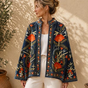 Könnte beinhalten: Dunkelblaue Jacke mit orangefarbener und grüner Blumenstickerei. Die Jacke hat einen Stehkragen und wird offen getragen. Das Model trägt ein weißes Oberteil und cremefarbene Hosen. Das Design der Jacke zeigt ein detailliertes Blumenmuster.