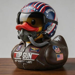 Può includere: Un'anatra di gomma gialla vestita da pilota, con un casco blu, bianco e rosso con la scritta MAVERICK, un giubbotto da aviatore marrone scuro con la scritta TOP GUN 40TH ANNIVERSARY 1986 2026, una maschera a ossigeno nera e un tubo grigio.