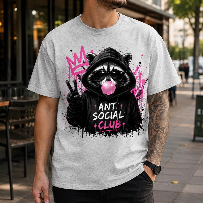 May include: A light gray t-shirt featuring a graphic of a raccoon wearing sunglasses and a black hoodie, blowing a bubblegum bubble. The raccoon is giving a peace sign. The words "ANTI SOCIAL CLUB" are printed below the raccoon in pink and white.