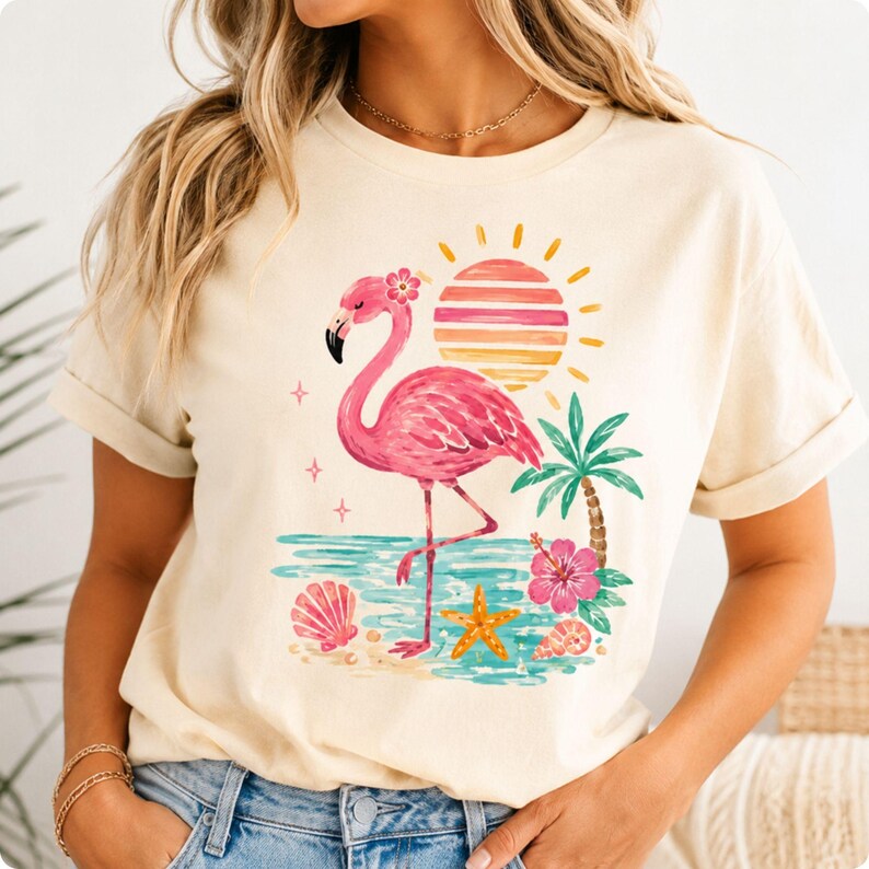 May include: A cream-colored t-shirt featuring a graphic print of a pink flamingo standing on a beach. The design includes a striped sun, a palm tree, a pink hibiscus flower, a seashell, and a starfish, all rendered in vibrant tropical colors.