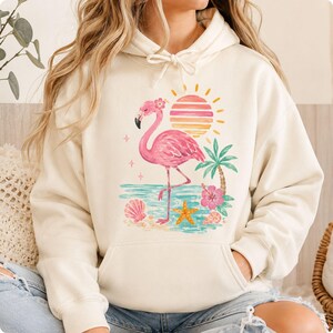 May include: A cream-colored hooded sweatshirt featuring a graphic print of a pink flamingo standing on a beach. The design includes a striped sun, a palm tree, a hibiscus flower, seashells, and a starfish, all rendered in soft pastel colors.