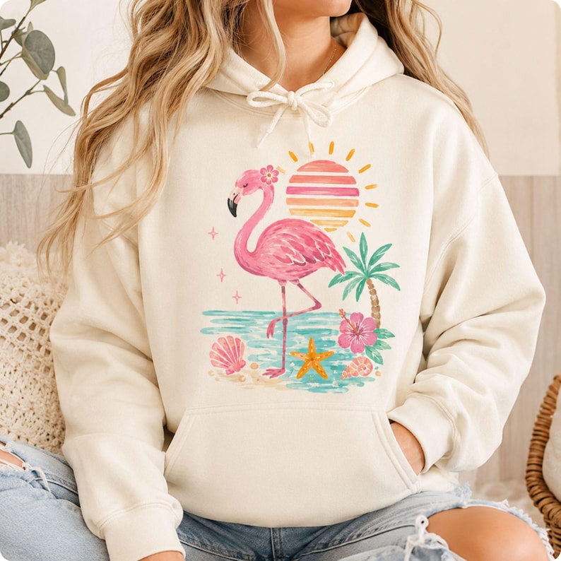 May include: A cream-colored hooded sweatshirt featuring a graphic print of a pink flamingo standing on a beach. The design includes a striped sun, a palm tree, a hibiscus flower, seashells, and a starfish, all rendered in soft pastel colors.