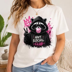 May include: White t-shirt featuring a raccoon wearing sunglasses and a black hoodie, blowing a pink bubblegum bubble. The raccoon is giving a peace sign. The words "ANTI SOCIAL CLUB" are printed on the hoodie in pink.