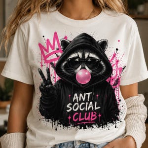 May include: White t-shirt featuring a raccoon wearing sunglasses and a black hoodie, blowing a pink bubble. The raccoon is giving a peace sign. The words "ANT SOCIAL CLUB" are printed on the hoodie in white and pink, with a pink crown and graffiti in the background.