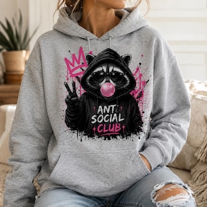 May include: A gray hoodie featuring a raccoon wearing sunglasses and a black hoodie with the text "ANTI SOCIAL CLUB". The raccoon is blowing a bubblegum bubble and giving the peace sign. Pink graffiti art is in the background.