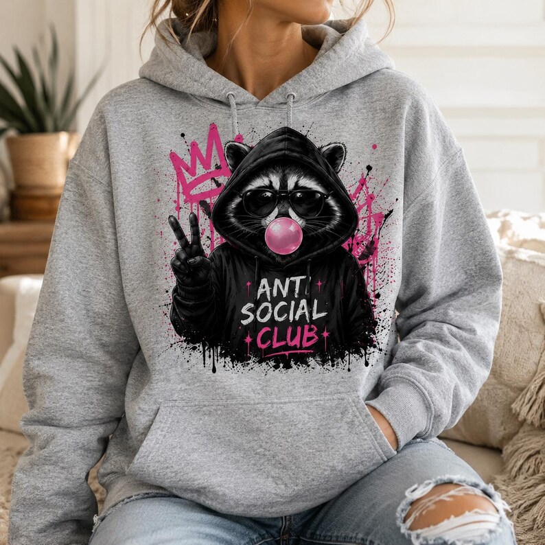 May include: A gray hoodie featuring a raccoon wearing sunglasses and a black hoodie with the text "ANTI SOCIAL CLUB". The raccoon is blowing a bubblegum bubble and giving the peace sign. Pink graffiti art is in the background.