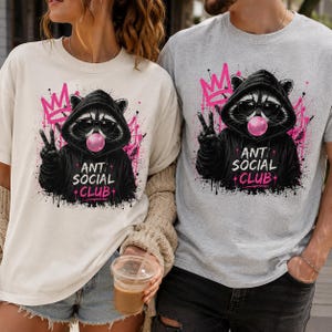 May include: Two t-shirts, one cream and one gray, featuring a raccoon wearing sunglasses and a hoodie, blowing a bubblegum bubble. The design includes a pink crown and the text "ANT SOCIAL + CLUB".