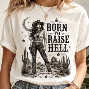 以下が含まれることがあります： オフホワイトのTシャツで、サボテンと三日月のある砂漠の風景の中にカウガールが描かれた黒いグラフィックプリントが施されています。テキストには「BORN TO RAISE HELL」および「wild from the start」と記されており、星や装飾的な線も含まれています。