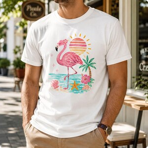 May include: A white short-sleeve t-shirt featuring a graphic print of a pink flamingo standing on a beach. The design includes a striped sun, a palm tree, a pink hibiscus flower, a starfish, and seashells in shades of pink, orange, and teal.