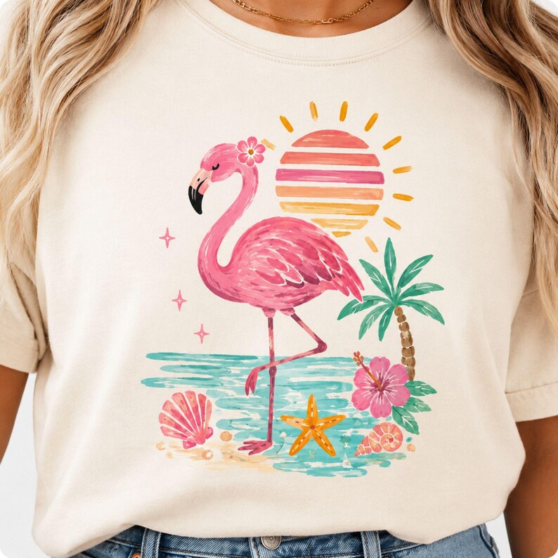May include: A cream-colored t-shirt featuring a graphic print of a pink flamingo standing on a beach. The design includes a striped sun, a palm tree, a hibiscus flower, a starfish, and seashells in shades of pink, orange, yellow, and teal.