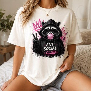 May include: Cream-colored t-shirt featuring a graphic of a raccoon wearing sunglasses and a black hoodie, blowing a bubblegum bubble. The hoodie has the text "ANTI SOCIAL CLUB" in white and pink. Pink graffiti-style accents surround the raccoon.