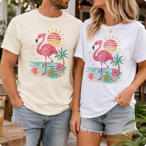 May include: Two short-sleeved t-shirts, one cream and one white, each featuring a graphic print of a pink flamingo standing on a beach. The design includes a striped sun, a palm tree, a hibiscus flower, a starfish, and seashells in warm colors.