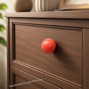 Può includere: Primo piano di un cassetto in legno con un pomello sferico rosso vibrante. Il cassetto è di un ricco colore marrone scuro e il pomello crea un contrasto sorprendente. La venatura del legno è visibile, aggiungendo consistenza al pezzo. Il pomello ha un diametro di circa 4 cm.