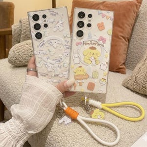 May include: Two clear phone cases with cartoon character designs. One case features a repeating pattern of white characters, while the other has a yellow dog character with the text "PomPomPurin" and "Happy Day". Attached are white and yellow wrist straps.