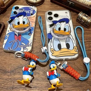 May include: Two clear smartphone cases featuring Donald Duck graphics. Each case includes a circular Donald Duck character pop-out grip and a blue lanyard with an orange braided section and a small Donald Duck figurine attached to the clip.