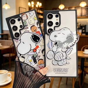 Könnte beinhalten: Zwei Handyhüllen mit Peanuts-Charakteren. Eine Hülle hat einen schwarzen Rand mit Snoopy, der ein rotes Objekt hält, umgeben von anderen Charakteren. Die andere Hülle hat eine transparente Rückseite mit Snoopy am Telefon. Das Wort "PEANUTS" ist sichtbar.