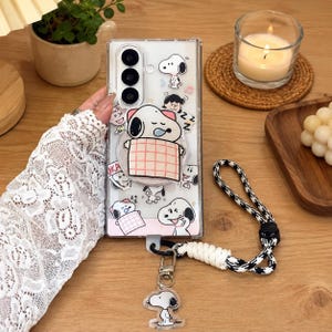 May include: A clear smartphone case featuring multiple Snoopy character illustrations, including a large Snoopy sleeping under a pink plaid blanket. The case includes a black and white houndstooth patterned wrist strap with a dangling Snoopy charm.