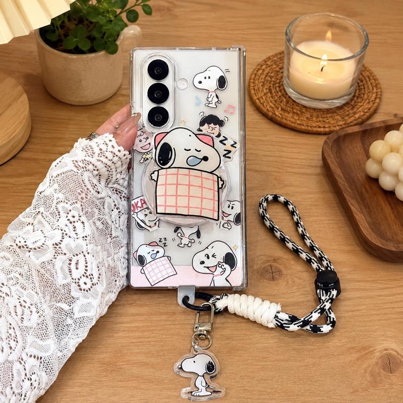 May include: A clear smartphone case featuring multiple Snoopy character illustrations, including a large Snoopy sleeping under a pink plaid blanket. The case includes a black and white houndstooth patterned wrist strap with a dangling Snoopy charm.