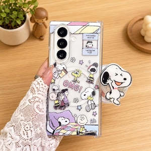 May include: A clear smartphone case featuring various Peanuts characters including Snoopy, Charlie Brown, and Woodstock. The design includes the text "OMG!", "Ah", and "Psychiatric Help 5&cent;". A circular magnetic ring is visible, and a Snoopy phone grip is attached.