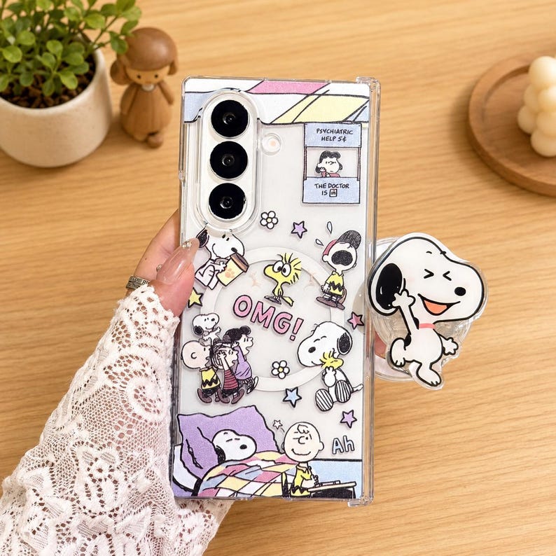 May include: A clear smartphone case featuring various Peanuts characters including Snoopy, Charlie Brown, and Woodstock. The design includes the text "OMG!", "Ah", and "Psychiatric Help 5&cent;". A circular magnetic ring is visible, and a Snoopy phone grip is attached.