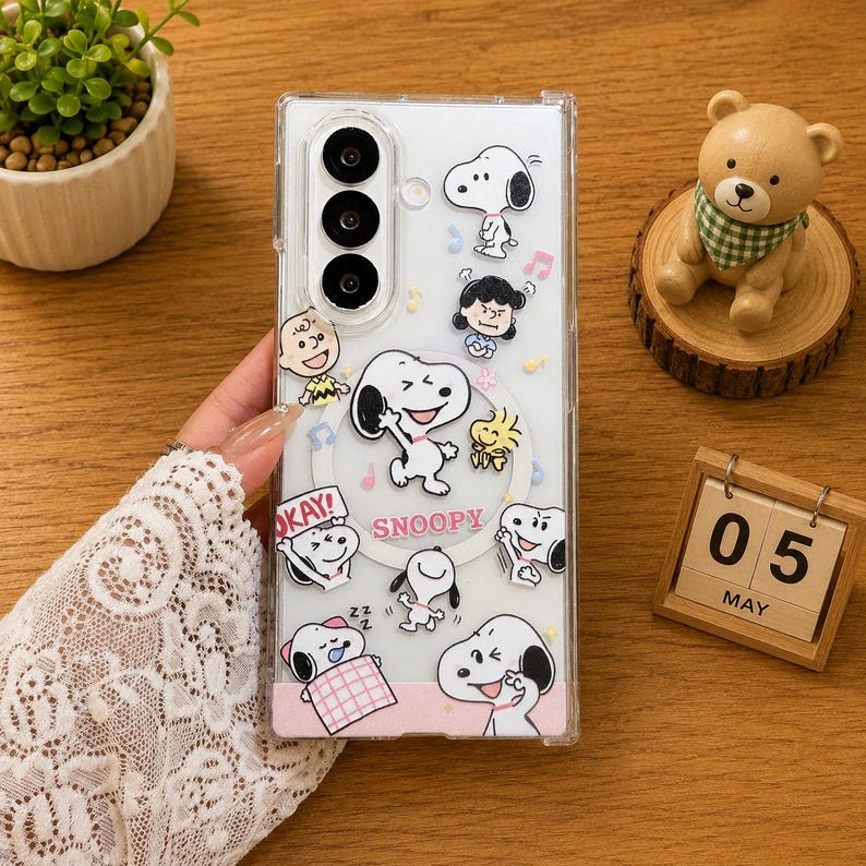 May include: A clear smartphone case featuring various Snoopy and Peanuts character graphics, including Snoopy, Woodstock, and Charlie Brown. The design includes the text SNOOPY in pink letters, musical notes, and small stars printed across the transparent surface.