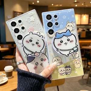 May include: Two clear phone cases with cartoon character designs. One case has a white and grey bear design, the other has a blue and white cat design. Both cases have a pop-up grip. The background is a blurred cafe setting.
