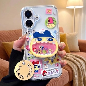 May include: A clear phone case featuring various colorful stickers, including a pink creature, a strawberry, and a Tamagotchi label. A circular, yellow, blue, and pink character-themed phone grip is attached, containing small floating charms.