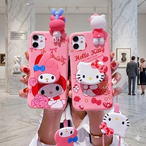以下が含まれることがあります： ハローキティとマイメロディのデザインが施された2つのピンク色の携帯電話ケース。各ケースには、上部に3Dキャラクターと、お揃いのチャームが付いています。ハローキティのケースには「Hello Kitty good night and sweet dreams」と書かれています。