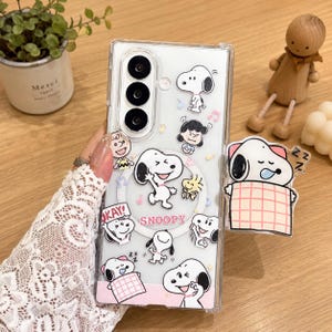 May include: A clear smartphone case featuring various Snoopy and Peanuts character illustrations, including Snoopy sleeping under a pink plaid blanket, dancing, and laughing. The case includes a circular magnetic ring and the text SNOOPY printed in red.
