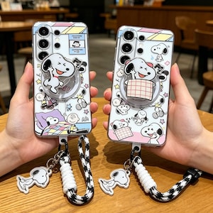 May include: Two clear phone cases with Snoopy and Peanuts characters. Each case has a Snoopy-shaped phone grip and a matching Snoopy charm on a lanyard. The cases have a white background with colourful accents.