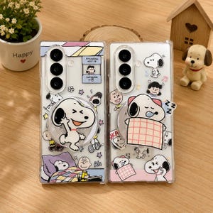 May include: Two clear rectangular phone cases featuring various Snoopy and Peanuts character graphics. The left case includes a circular Snoopy pop-out grip and a psychiatric help booth illustration. The right case features a sleeping Snoopy grip.
