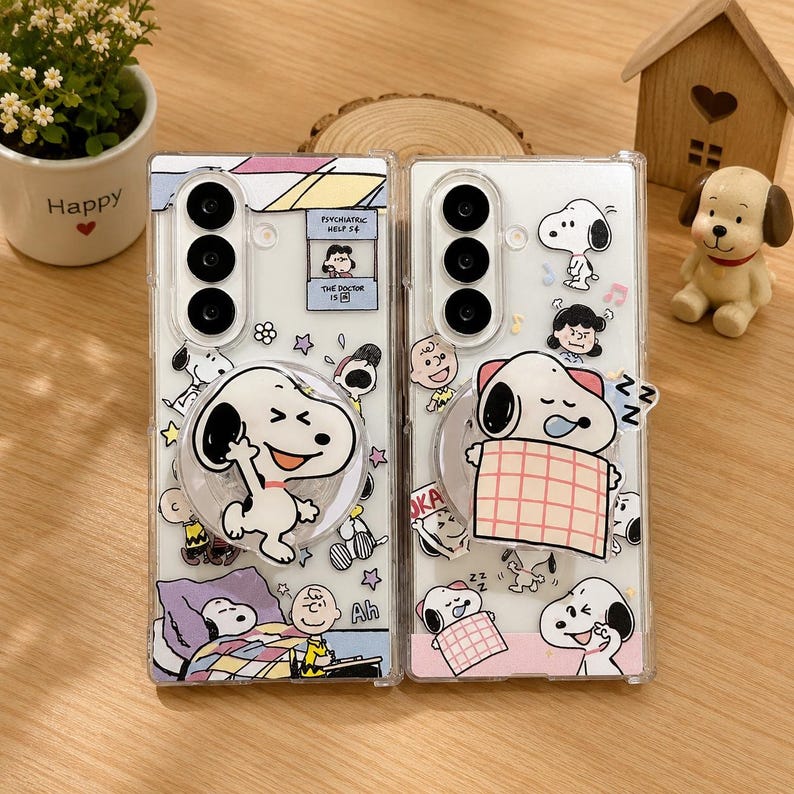 May include: Two clear rectangular phone cases featuring various Snoopy and Peanuts character graphics. The left case includes a circular Snoopy pop-out grip and a psychiatric help booth illustration. The right case features a sleeping Snoopy grip.