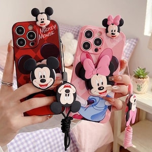 Könnte beinhalten: Zwei Handyhüllen mit Mickey- und Minnie-Mouse-Designs. Eine Hülle ist rot mit einem Mickey-Mouse-Muster und dem Text "Mickey Mouse". Die andere ist rosa mit einem Minnie-Mouse-Design. Beide Hüllen haben 3D-Zeichenbefestigungen und Handschlaufen.