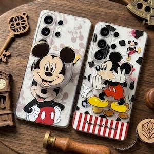Può includere: Due custodie trasparenti per telefono con disegni di Topolino. Una custodia ha una grande faccia di Topolino con un'impugnatura pop-up. L'altra custodia ha un'illustrazione a figura intera di Topolino con il testo "Mouse and Friends" su un bordo a righe rosse e bianche.